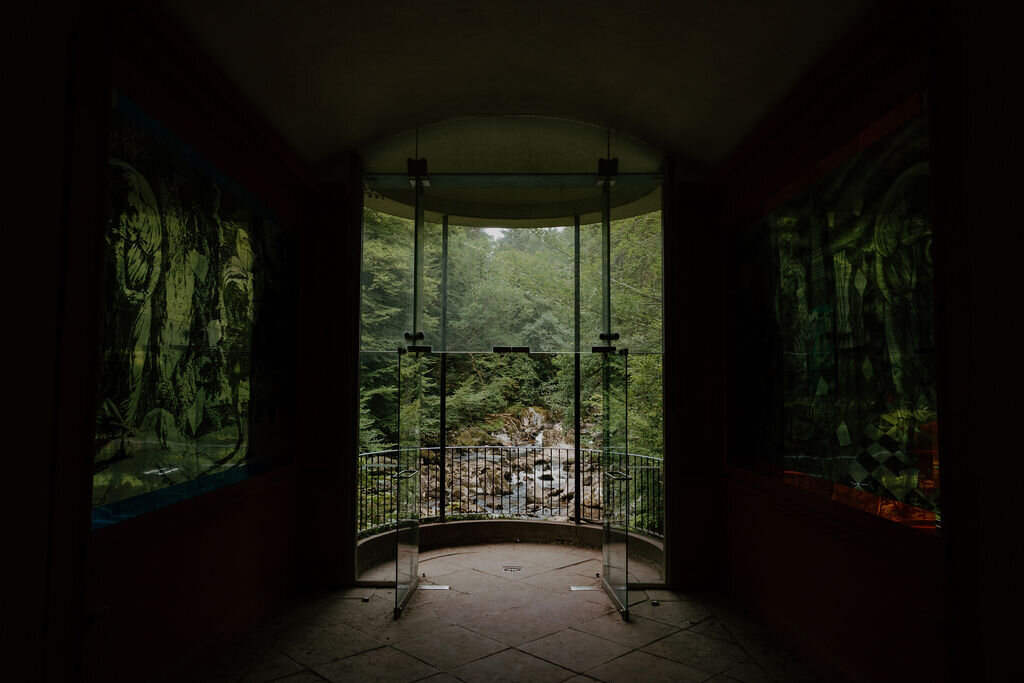 Glass doors of Ossian's Hall frame a view over the wooded river gorge at The Hermitage, Dunkeld, flanked by painted wall panels.