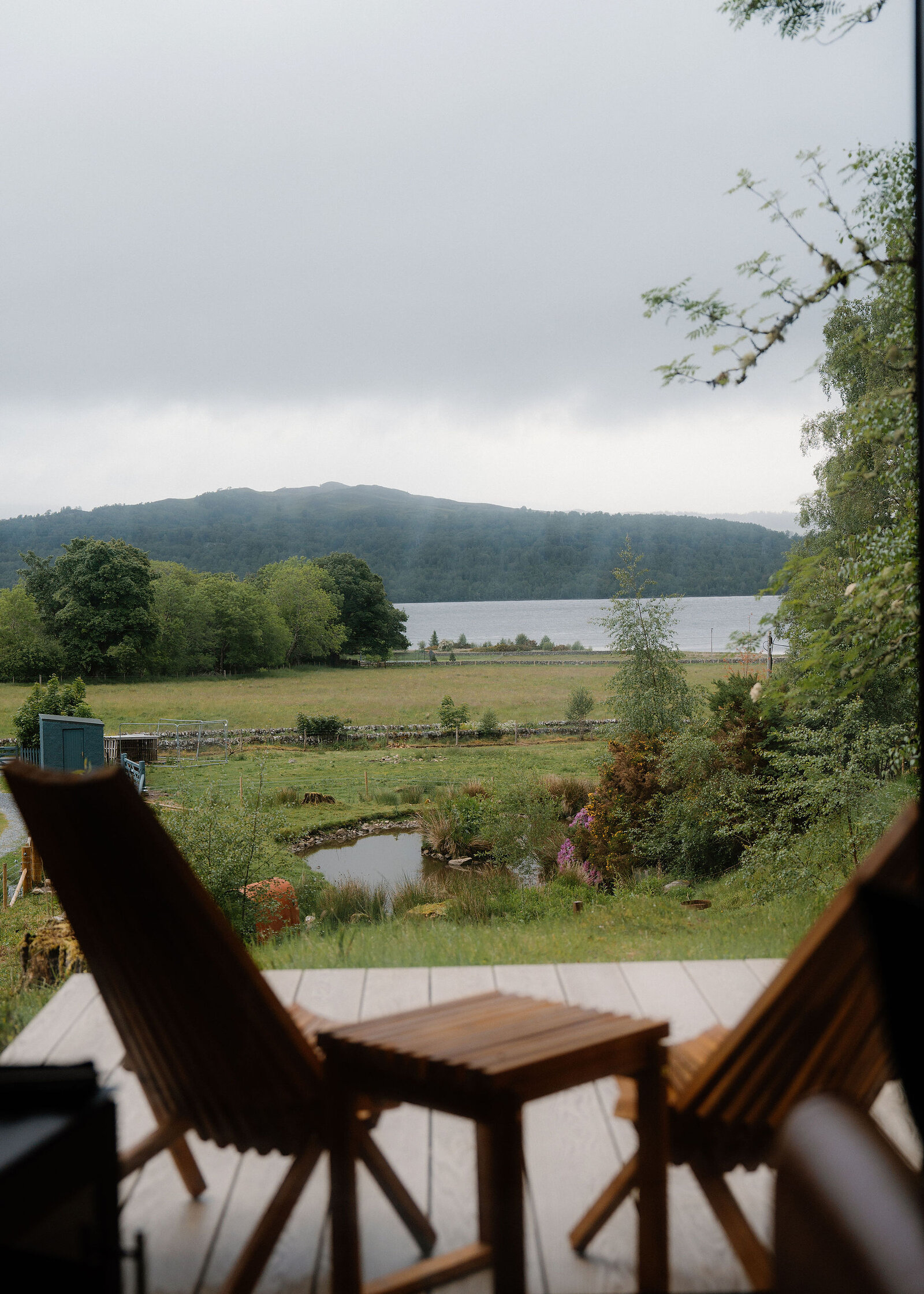 Two slatted wooden chairs on the deck of Loch face across rough fields and a pond to Loch Rannoch under grey cloud.