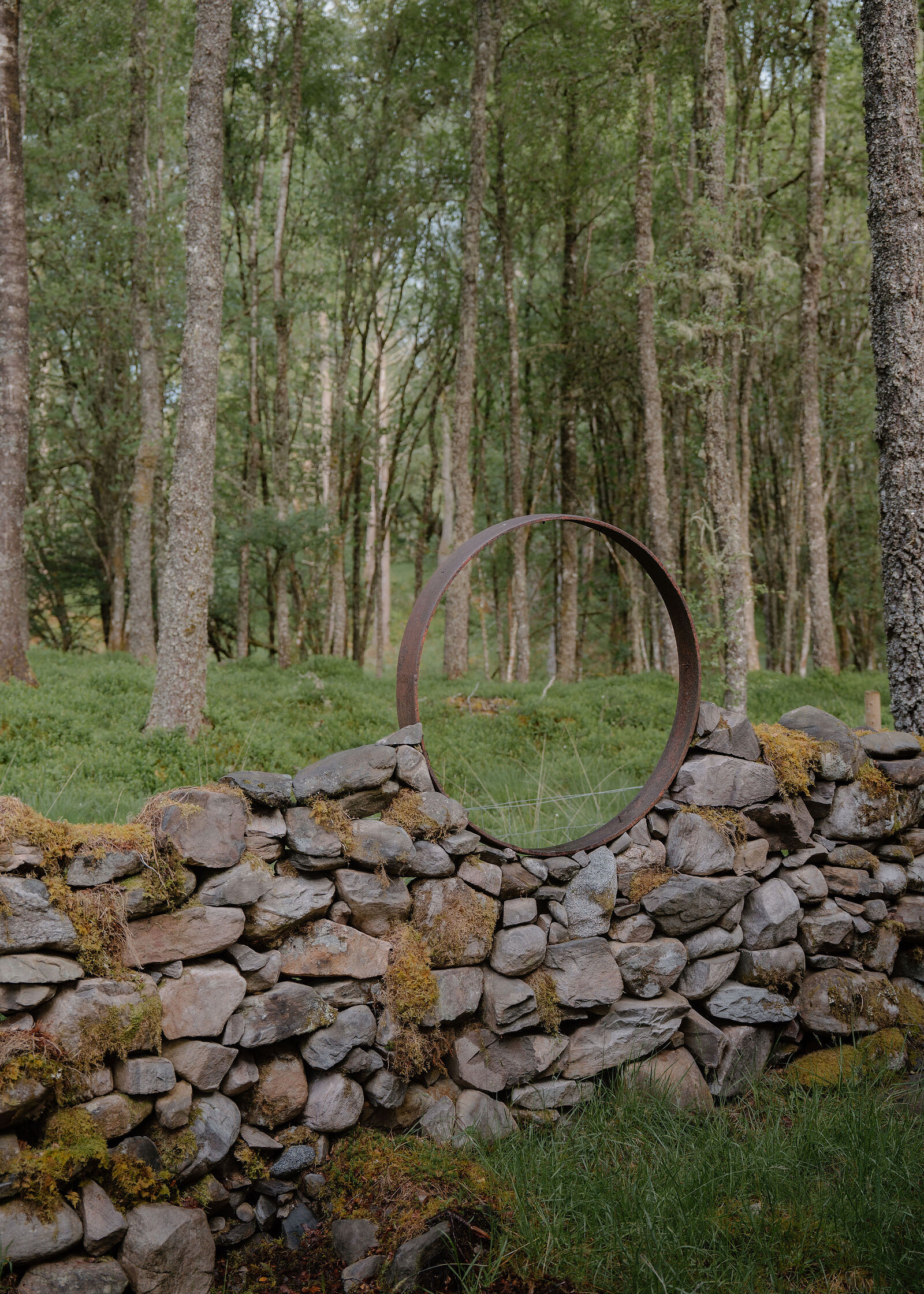 A rusted steel ring set into a mossy drystone dyke frames the birch wood beyond, the moon gate outside Rannoch.