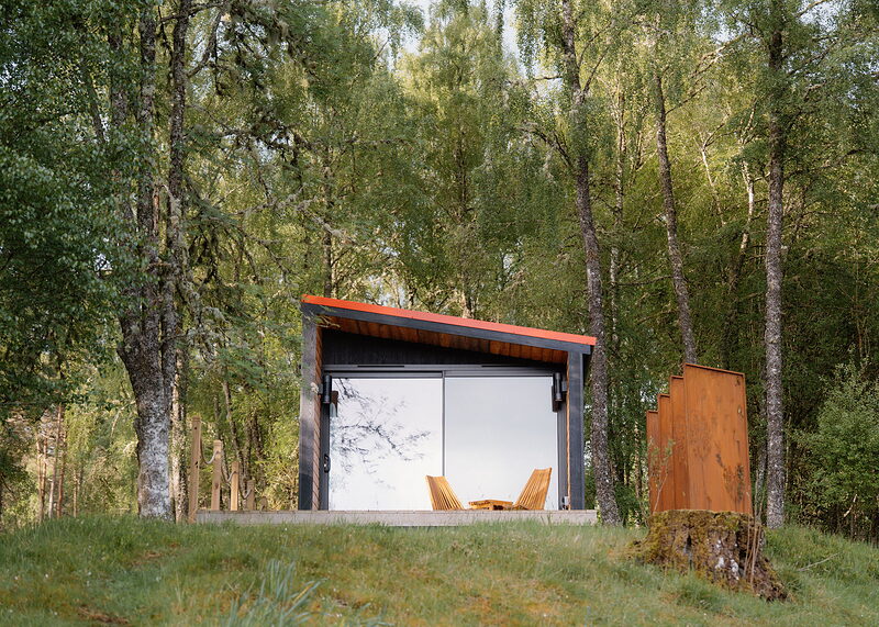 A landscape view of Loch on the crest of its grassy bank, the glass front reflecting the birches and a rusted screen by an old stump.