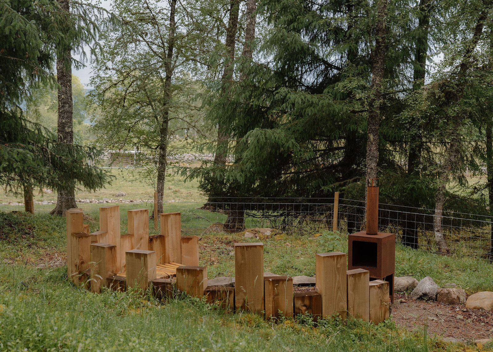 A landscape view of the fire pit's seats of upright timbers of different heights and its rusted stove, a fence and spruce behind.