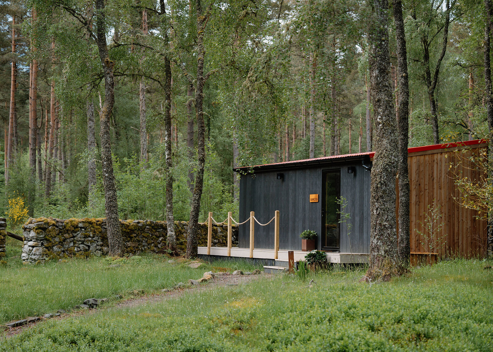 Rannoch from across the clearing: a black cabin with a larch end, a rope-railed deck and a mossy drystone dyke, tall pines behind.