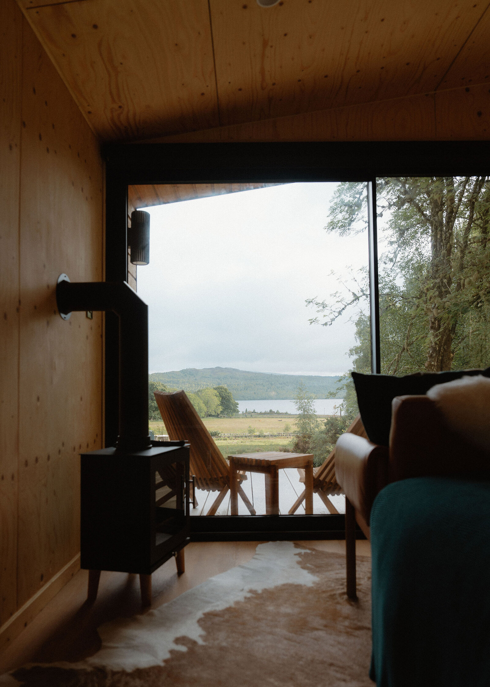 Inside Loch, the black stove stands by the glass front, the deck chairs and Loch Rannoch beyond and a cowhide on the floor.