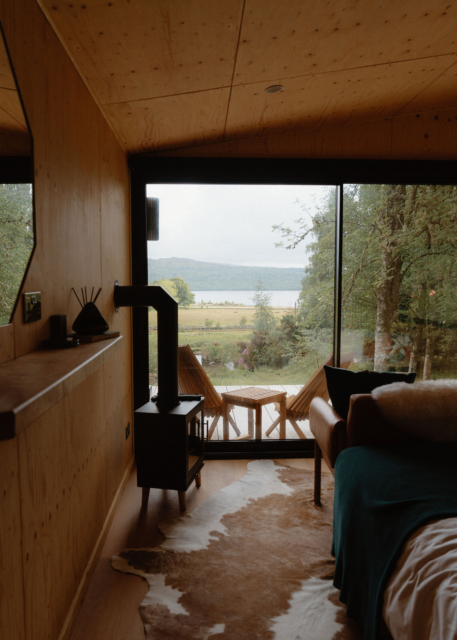 Down the length of Loch to its glass front: a shelf, the black stove, a cowhide rug and the loch beyond the deck.