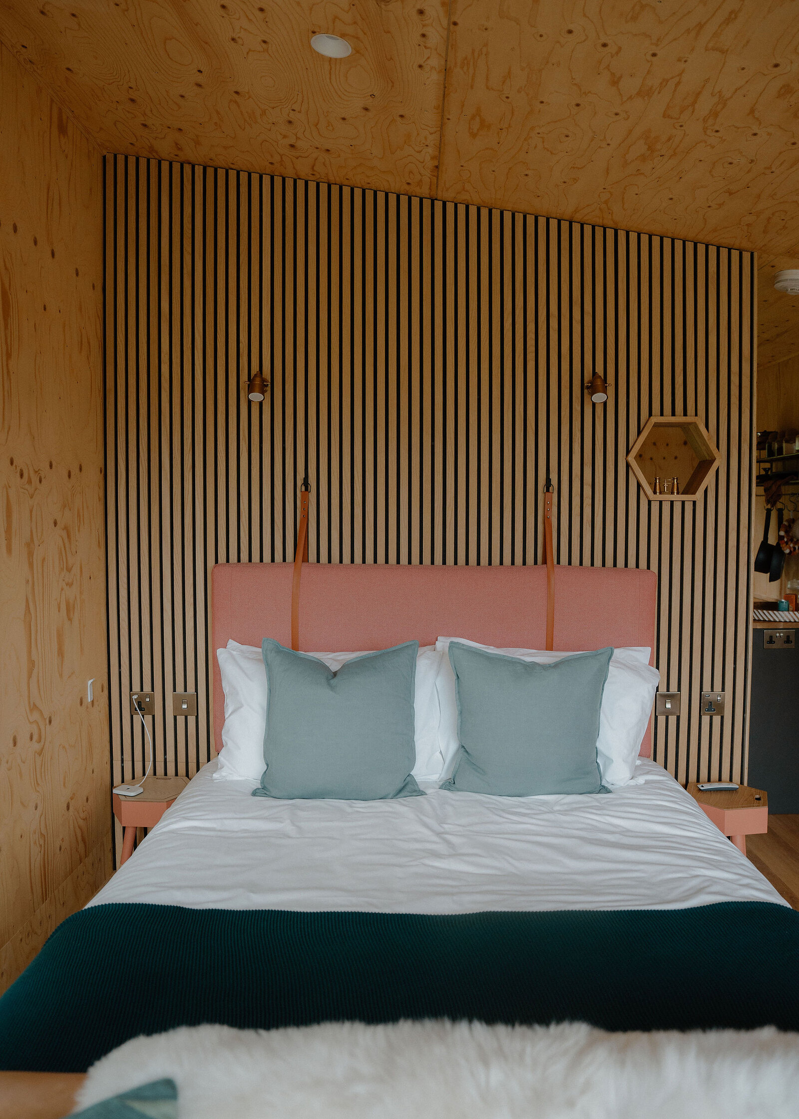 Loch's bed against slatted timber with a coral headboard on leather straps, teal cushions, a hexagonal mirror and a sheepskin at its foot.