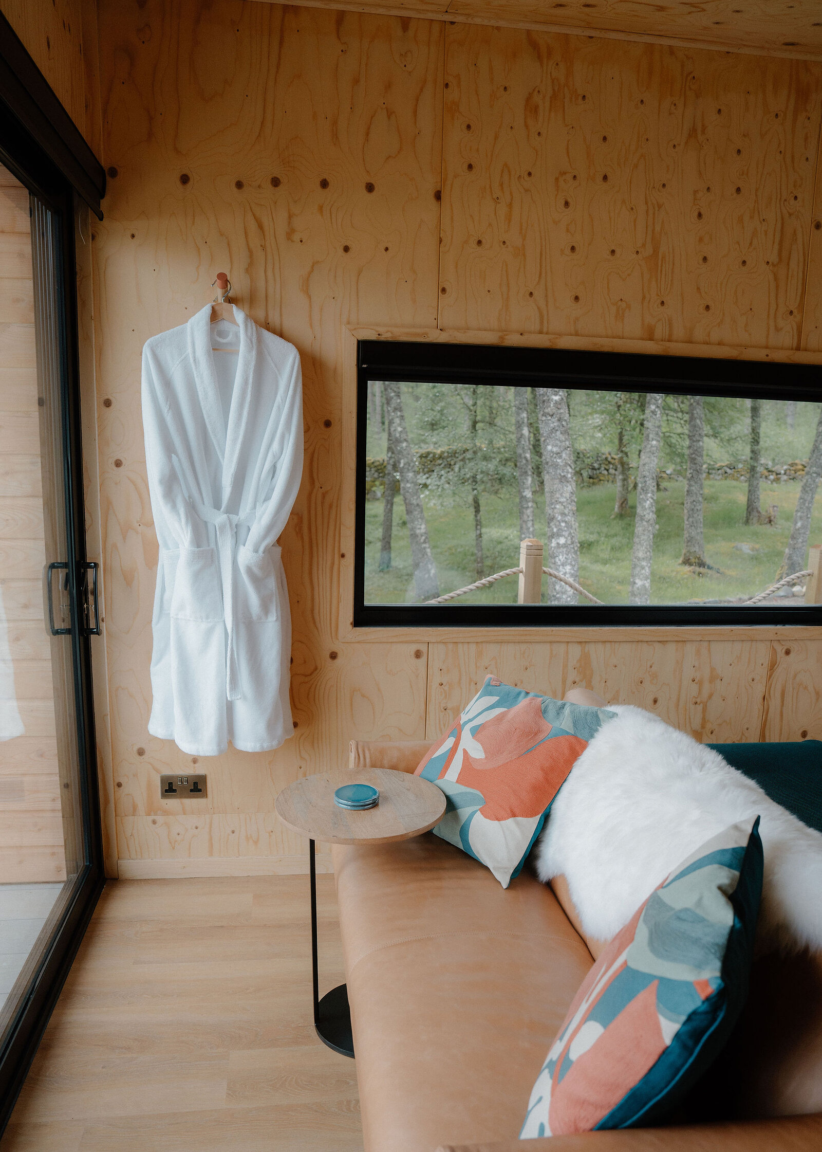 A tan leather love seat with a sheepskin and coral and teal cushions below a long window onto the woods, a robe by the door.