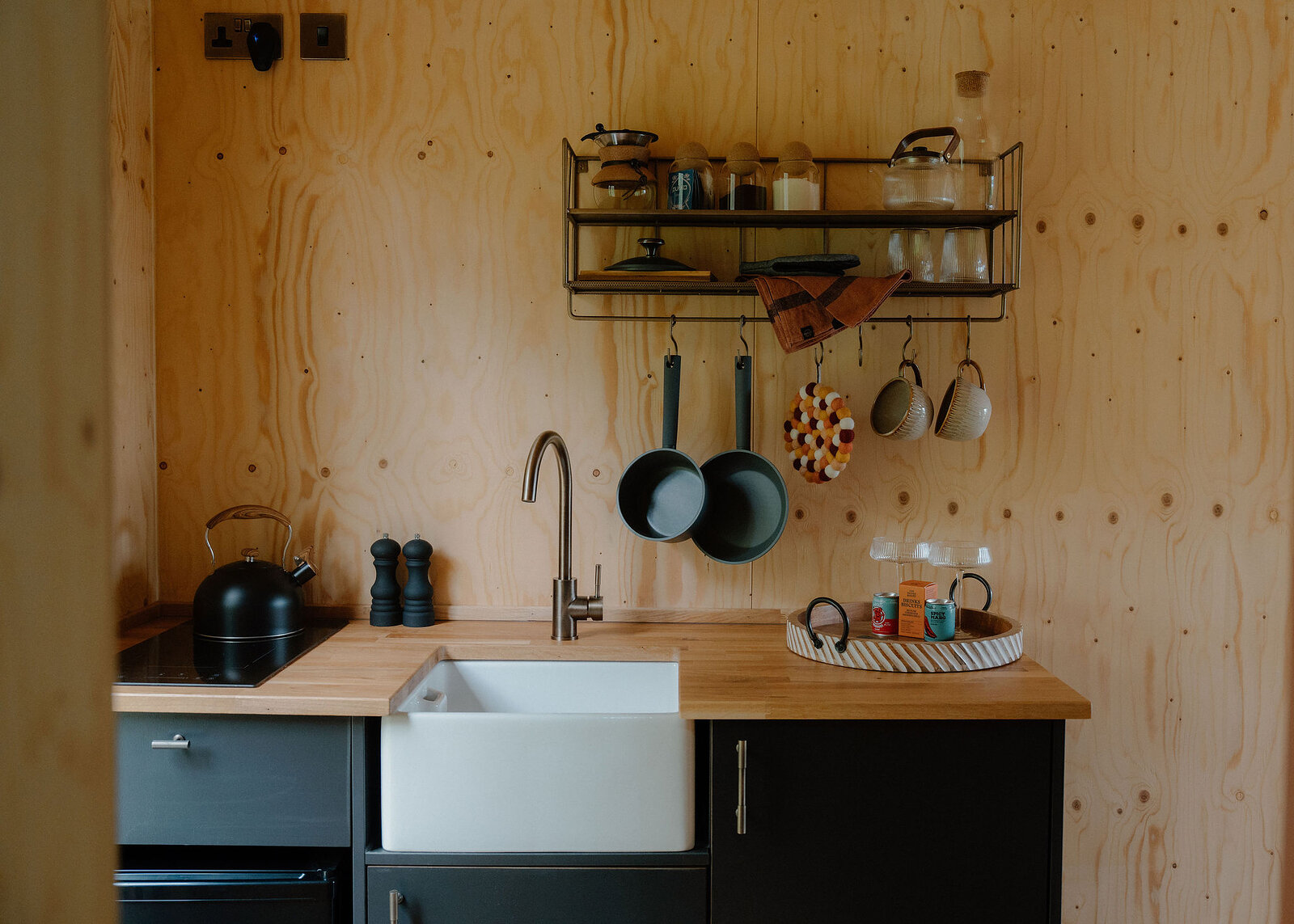 A landscape view of the kitchenette: kettle on the induction hob, pepper mills, sink, hanging pans and a tray of drinks.