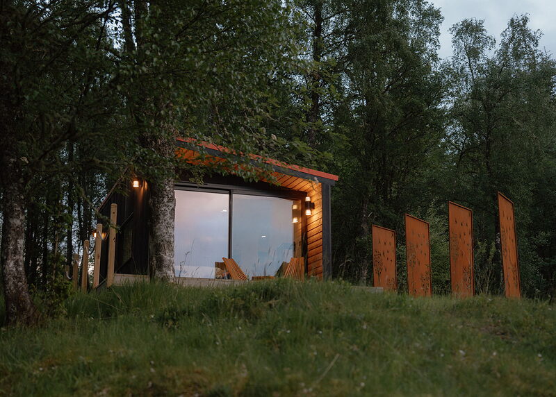 A landscape view of Loch at dusk, lamps glowing under the roof and a line of rusted steel screens beside it on the knoll.