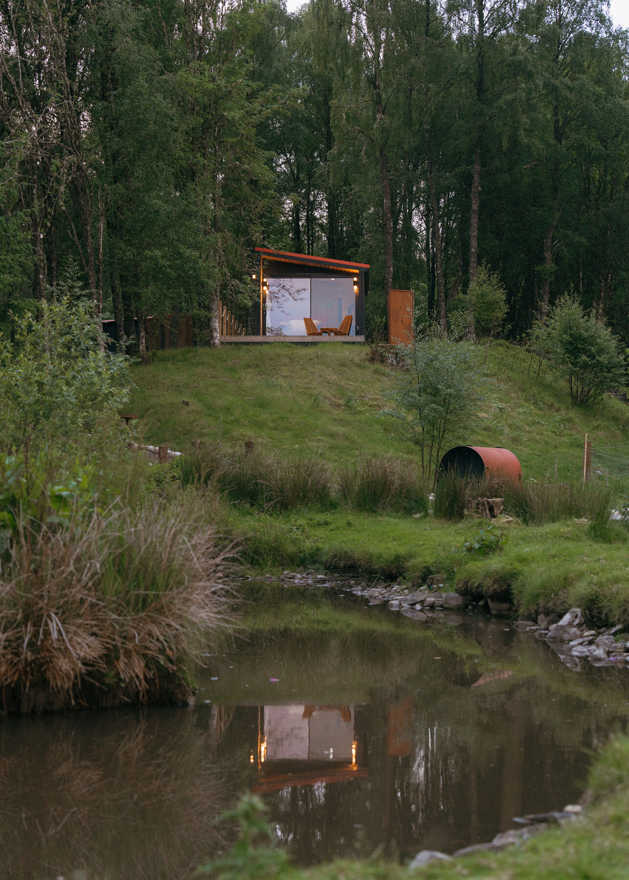 Loch lit on its knoll at dusk, reflected in the pond below, the barrel shelter on the bank between.