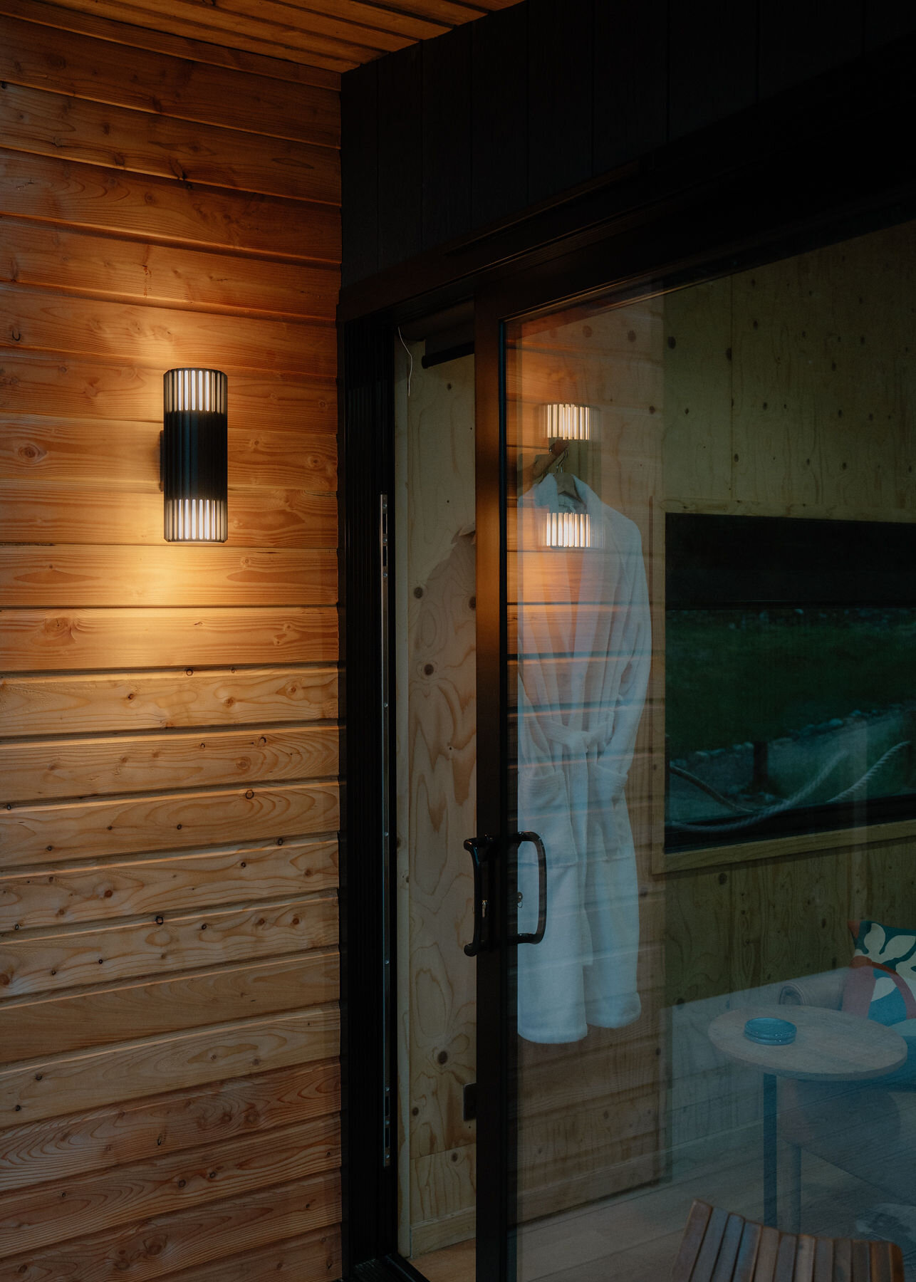 A wall lamp glows on the larch by Loch's glass door at dusk, a robe hanging inside.
