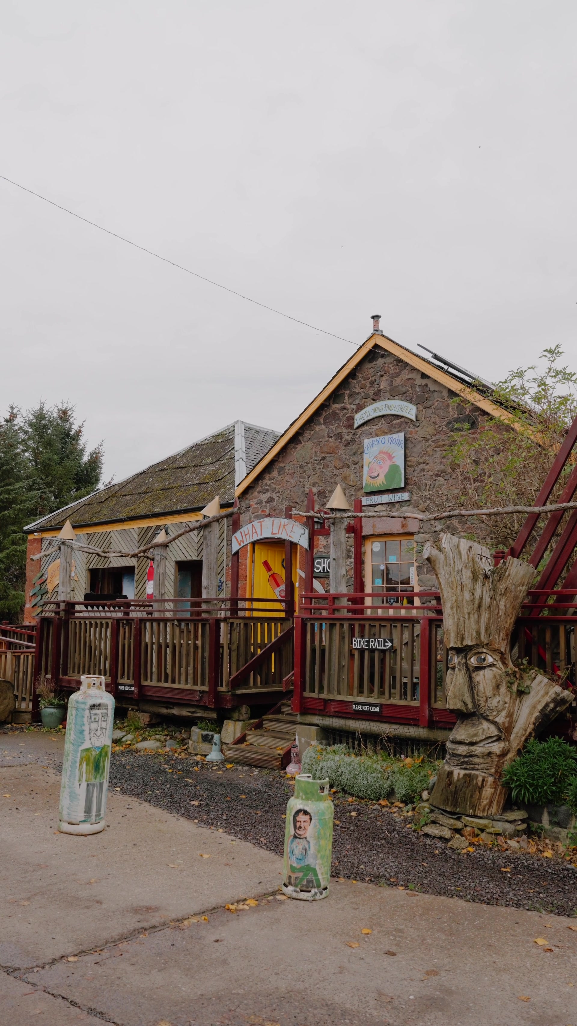 Cairn O' Mhor's stone fruit-wine shop, its 'What Like?' sign over a yellow door and a carved wooden face beside the deck.