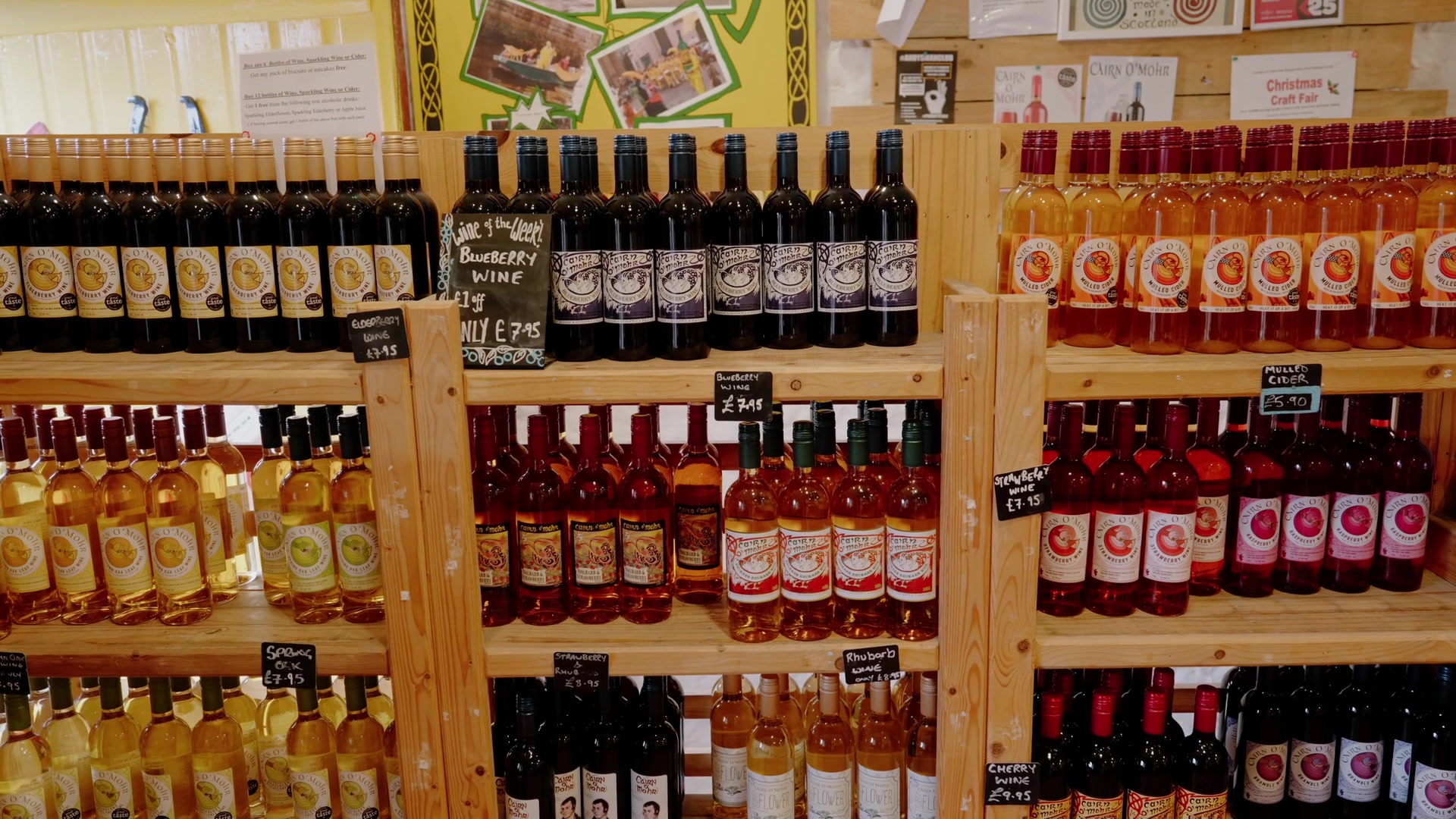 Shelves of Cairn O' Mhor fruit wines and mulled cider, handwritten price tags beneath a display of photographs.