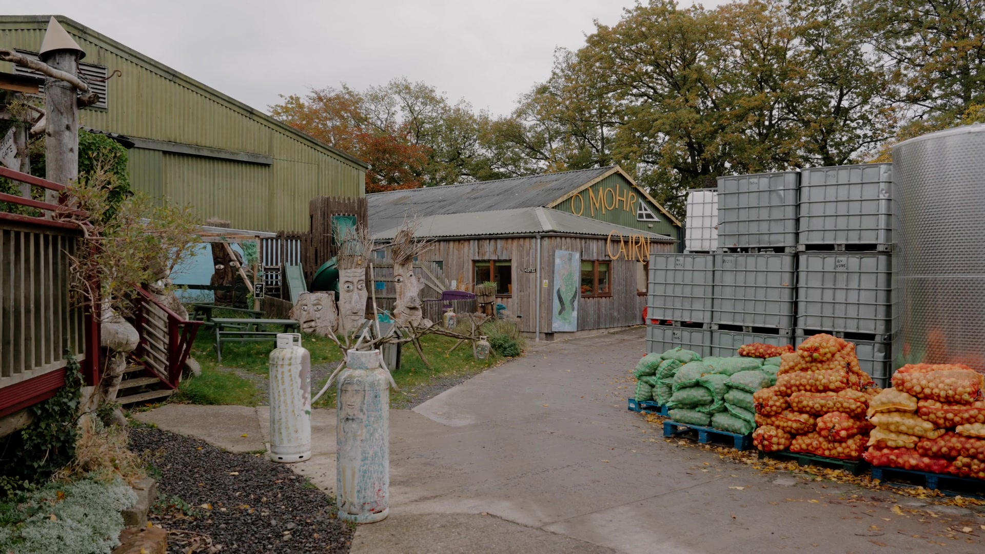 A timber building signed "O Mohr Cairn", carved wooden totem faces and painted gas cylinders beside a stack of bagged produce.