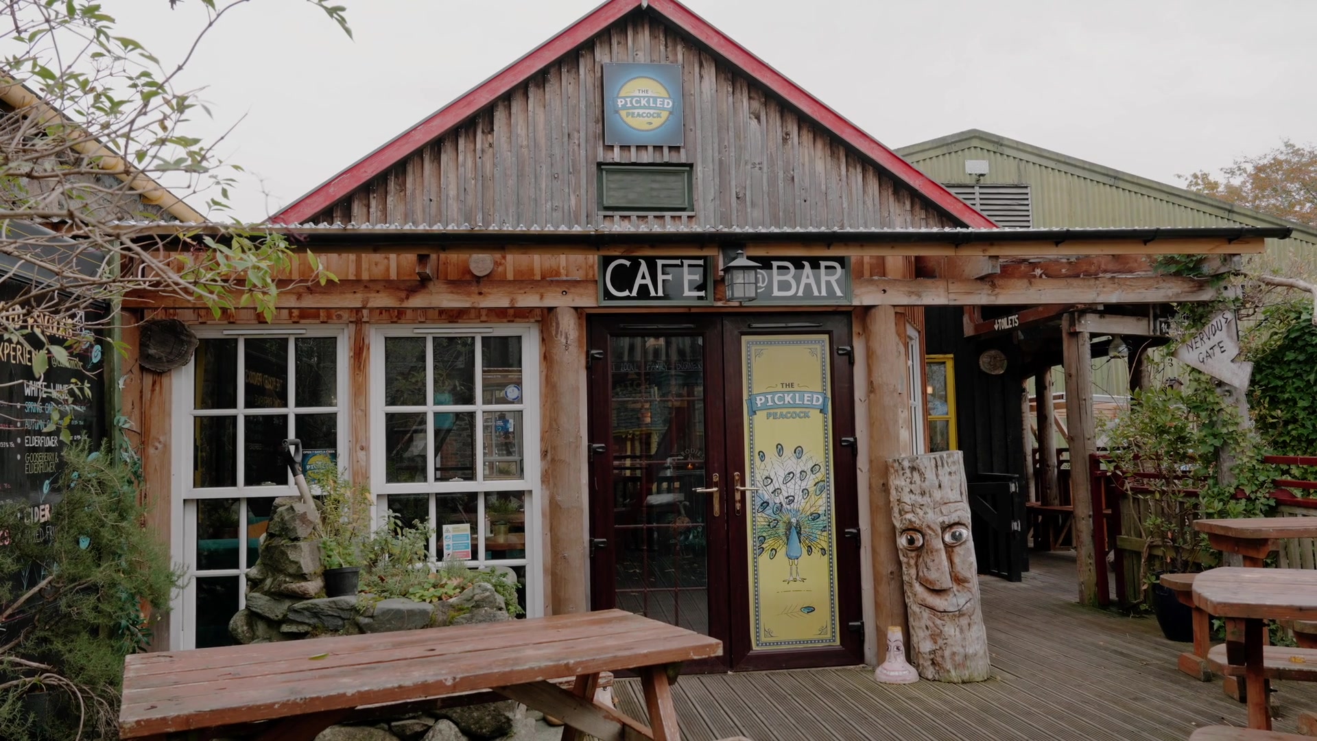 "The Pickled Peacock" sign above a "Cafe Bar" entrance, a peacock painted on the glass door and a carved wooden totem beside it.