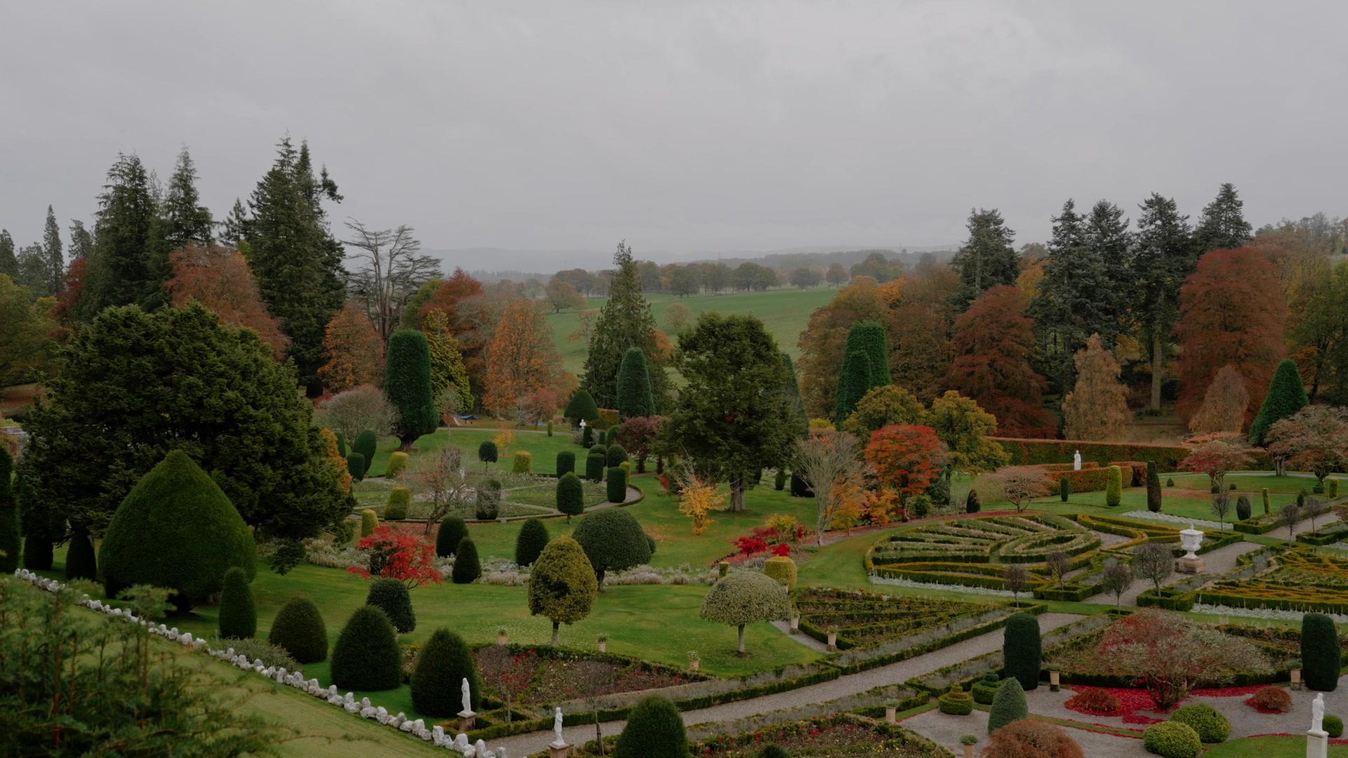 A turreted stone castle overlooks a reflecting pond, a stone bridge and topiary garden framed by dark conifers.