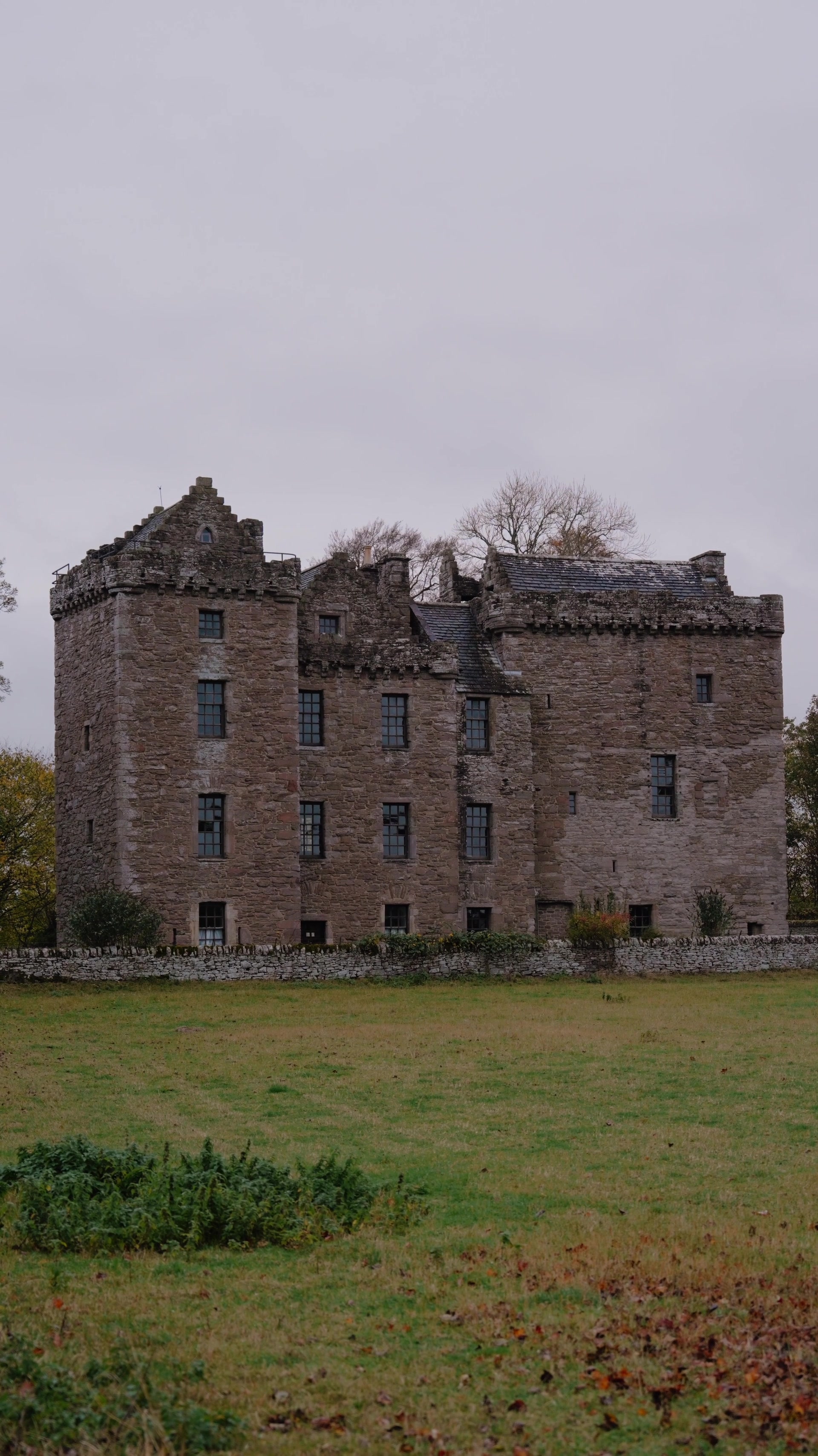 A ruined stone tower house with crow-stepped gables and battlements stands behind a dry-stone wall in an open field.