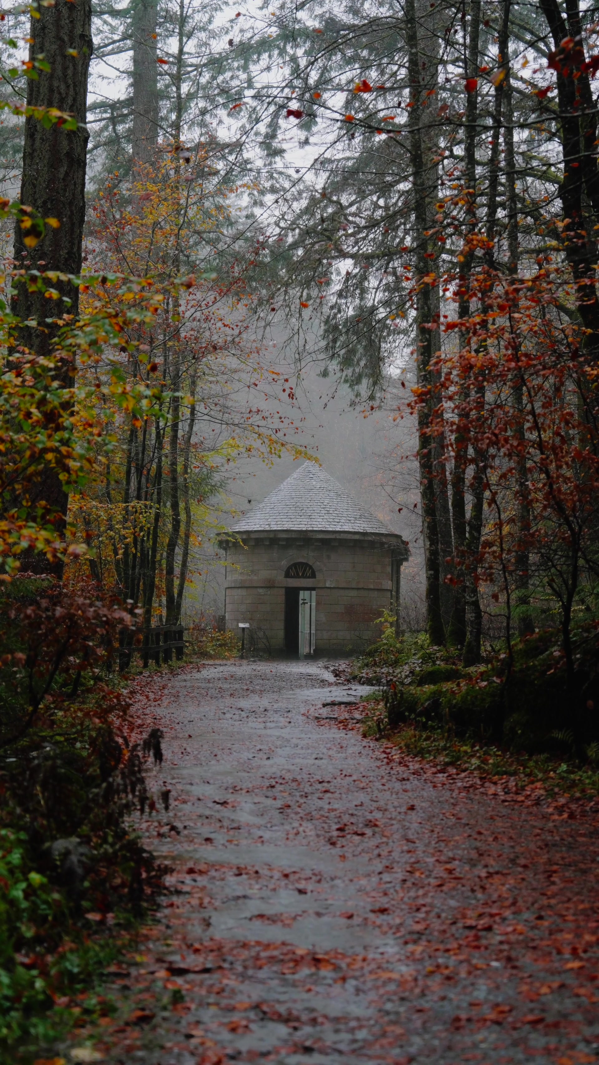 a round stone pavilion with a conical roof and MVM carved above its door, standing at the end of a leaf-strewn woodland path