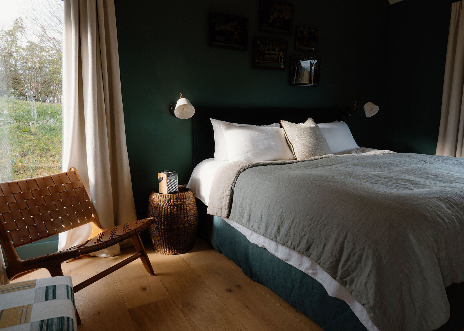 A landscape view of the green bedroom: the bed in soft linen, the leather chair and the window onto a bank of grass and trees.