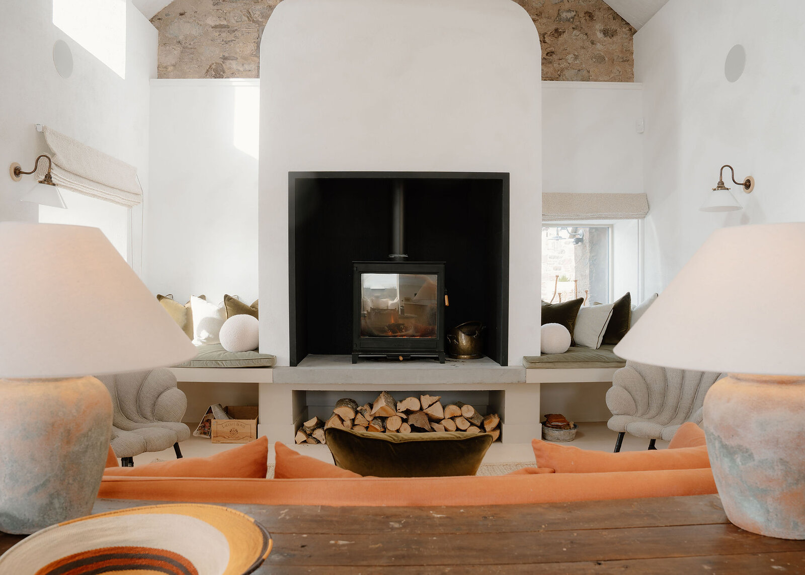 A landscape view of the stove and its log store between two window seats of cushions, big lamps on the table in front.