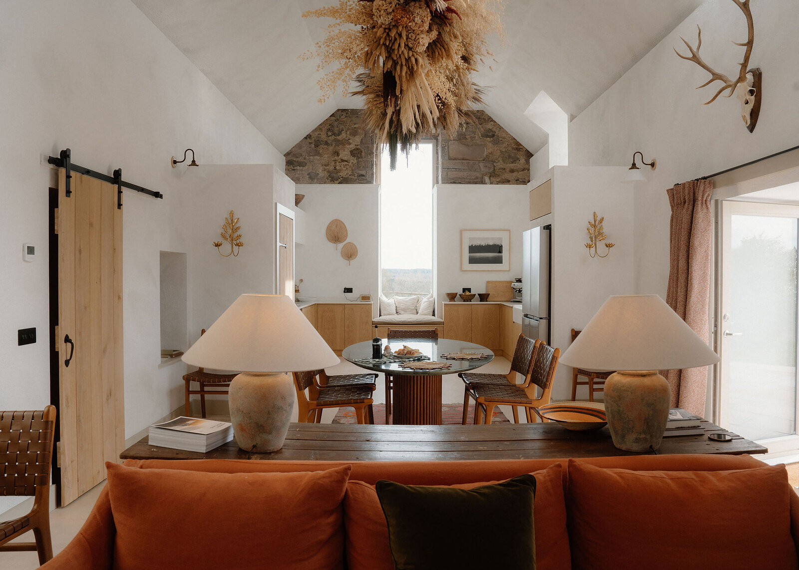 A landscape view of the whole main room: rust sofa, long table and lamps, the round dining table, the kitchen and stags' antlers under the vaulted ceiling.