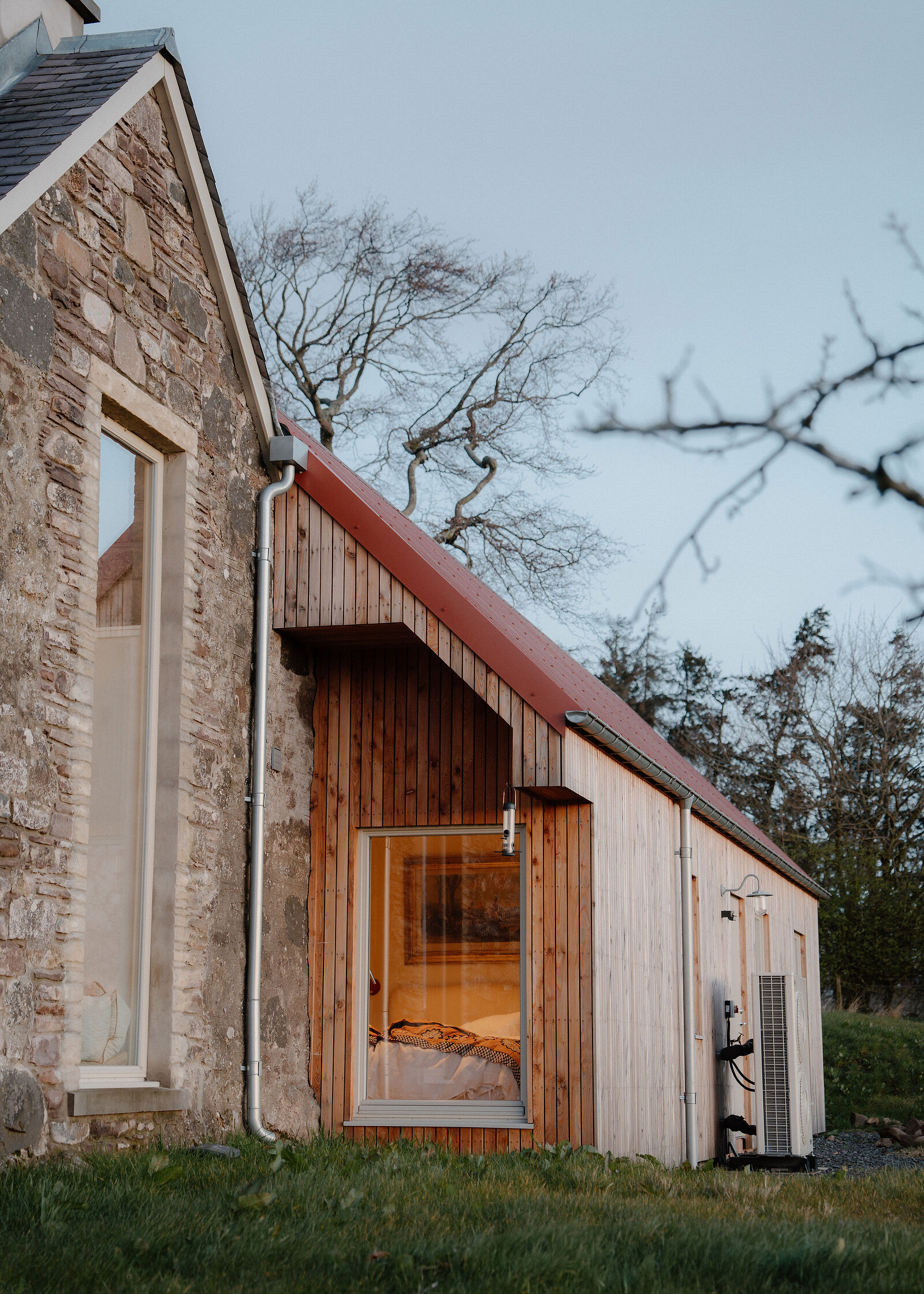 Where the old stone gable meets the larch-clad bedroom wing, a lit bedroom behind its big window at dusk.