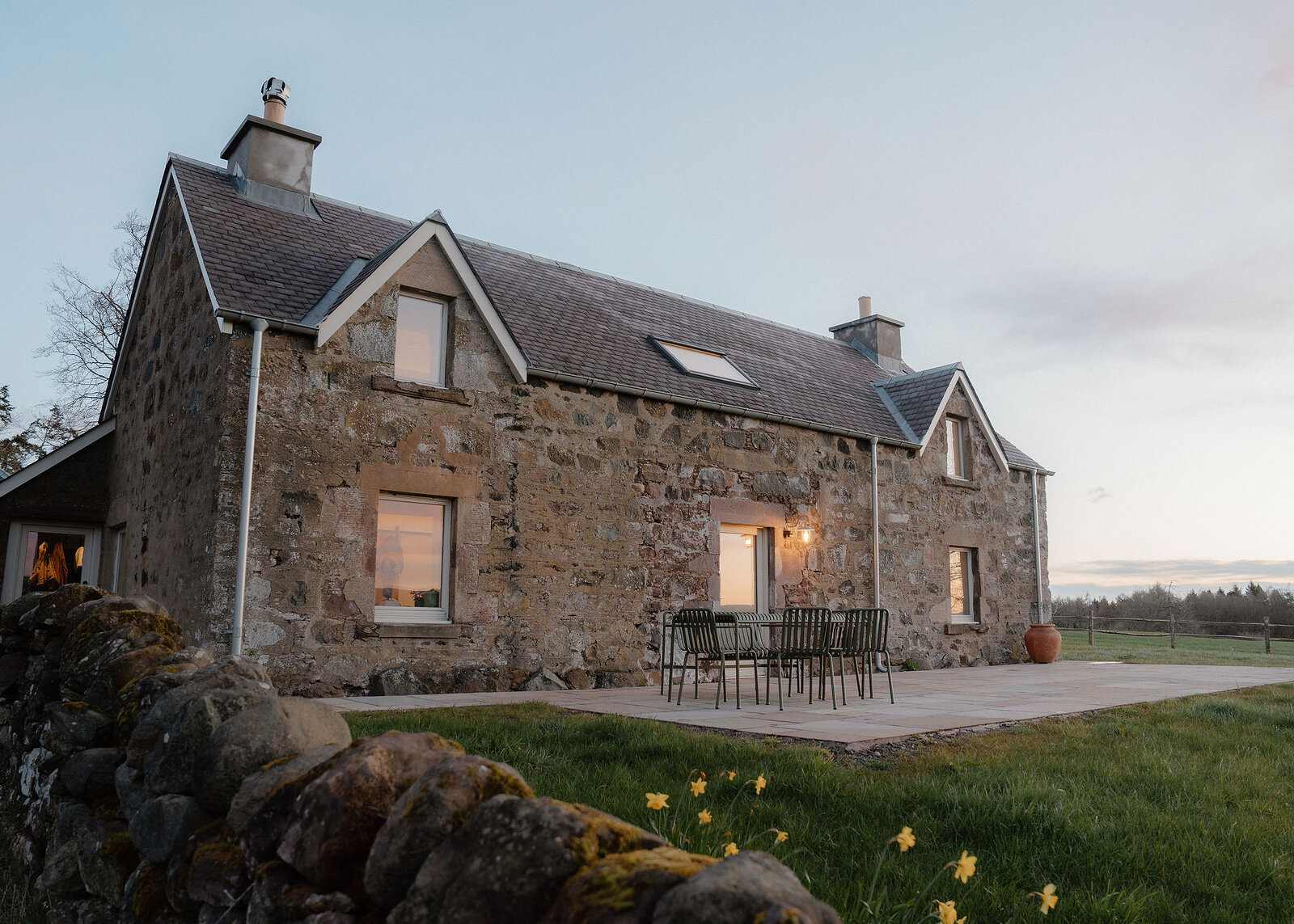 A landscape view of the whole stone house at dusk, its lit windows, terrace and a terracotta pot, the mossy dyke in front.