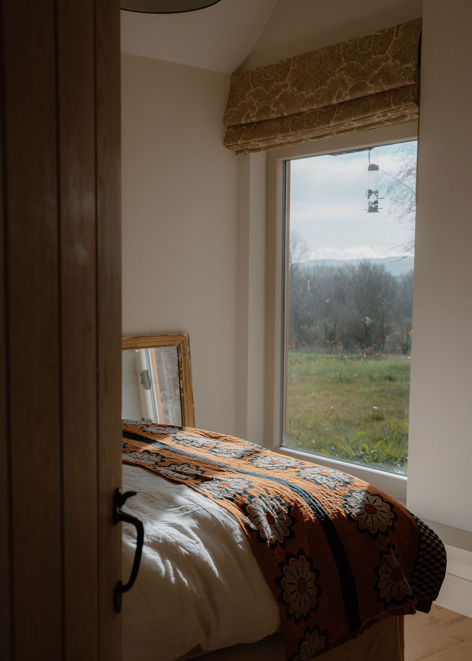 Past an oak door, a bed with a patterned orange throw by a window onto the fields and a bird feeder.