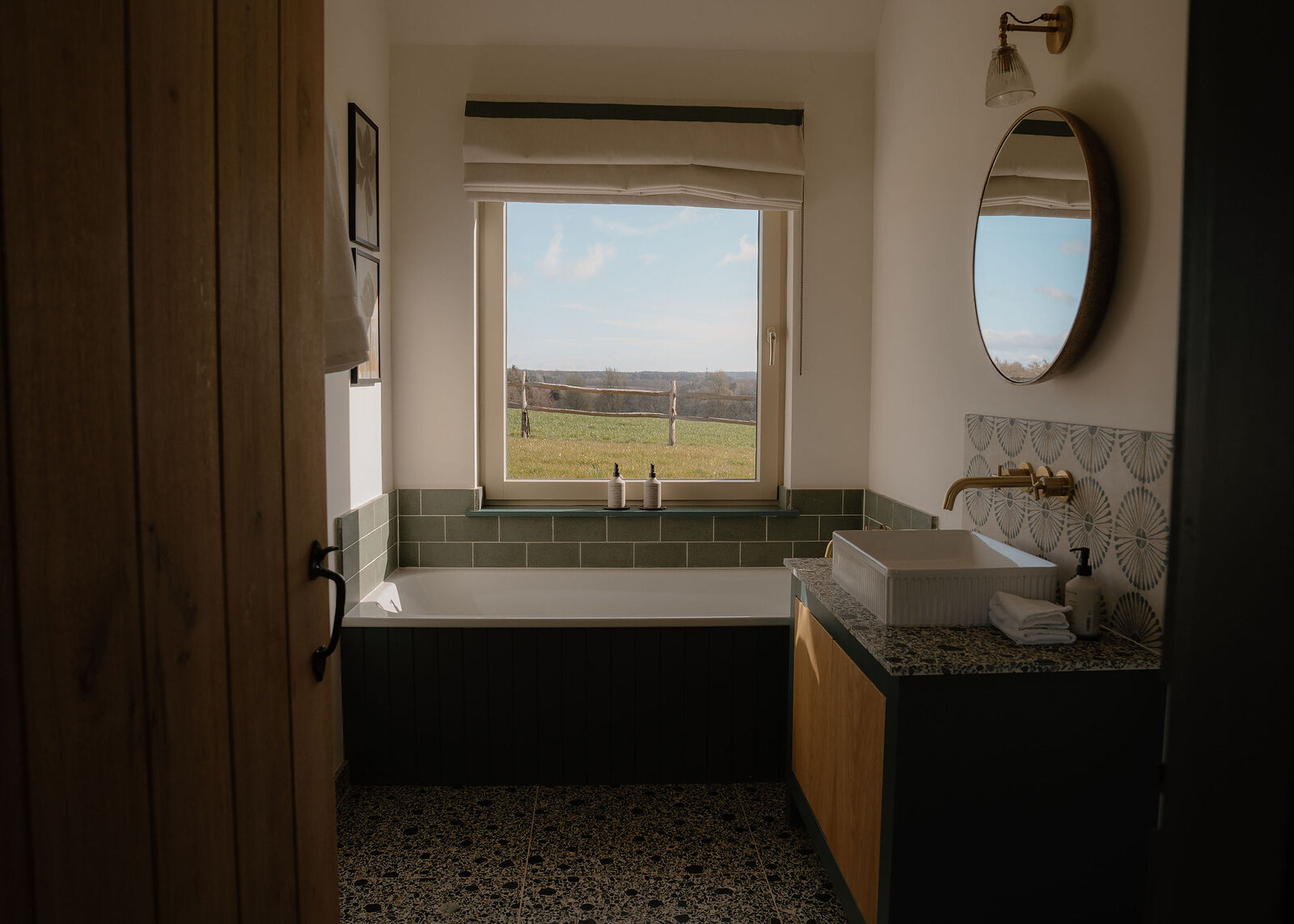 The en-suite through its oak door: a bath under the window, a basin on an oak cabinet, a round mirror and a terrazzo floor.