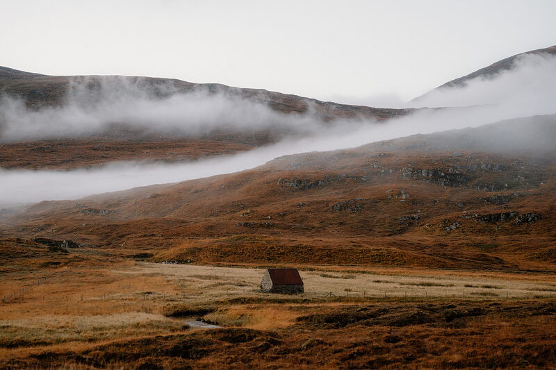 A small stone bothy with a red roof sits in a golden glen beneath mist-covered mountains.