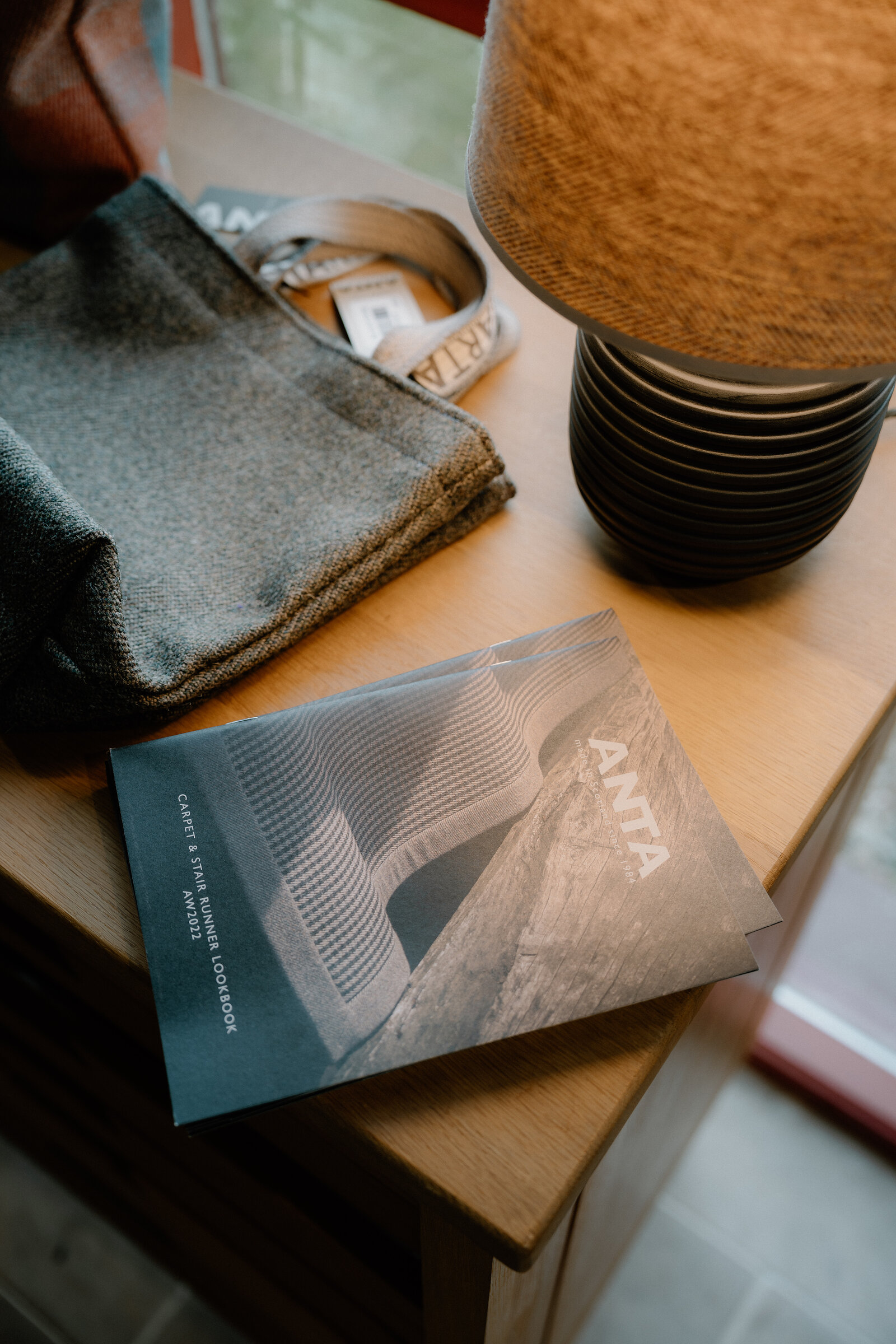 An ANTA cookbook lies beside a folded scarf and a woven lampshade on a wooden table.