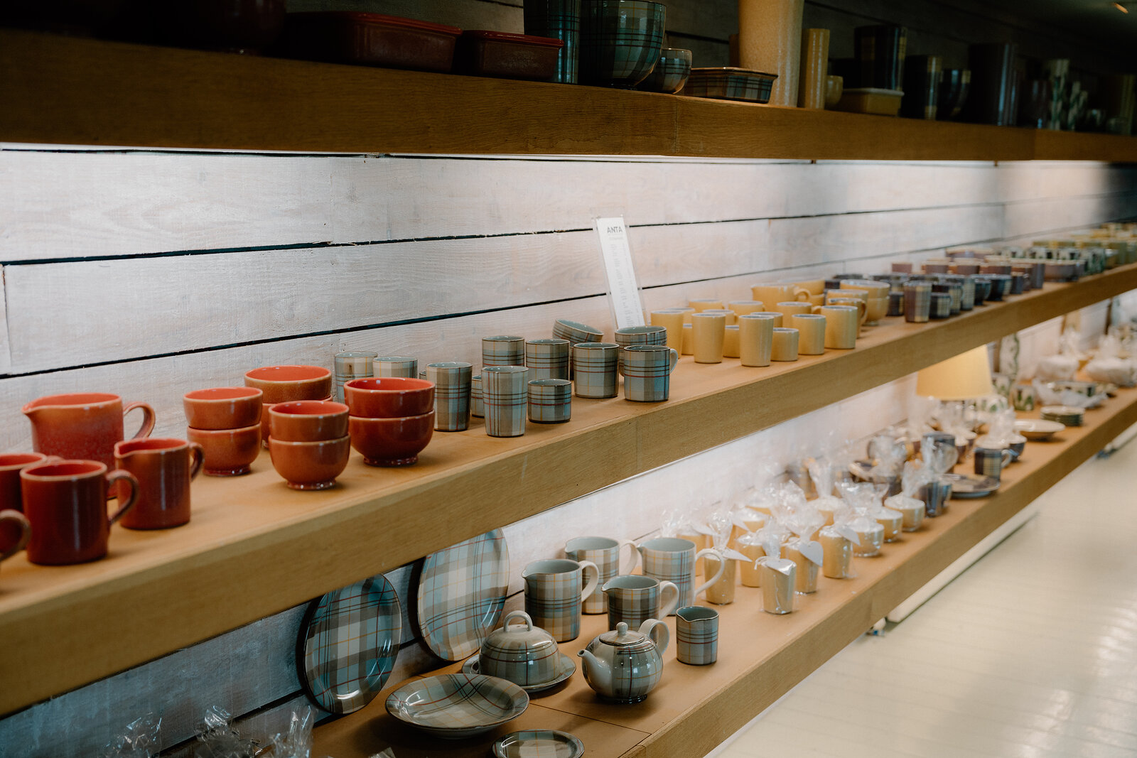 Glazed mugs, bowls and plates in red, blue and check patterns line wooden shelves at ANTA.