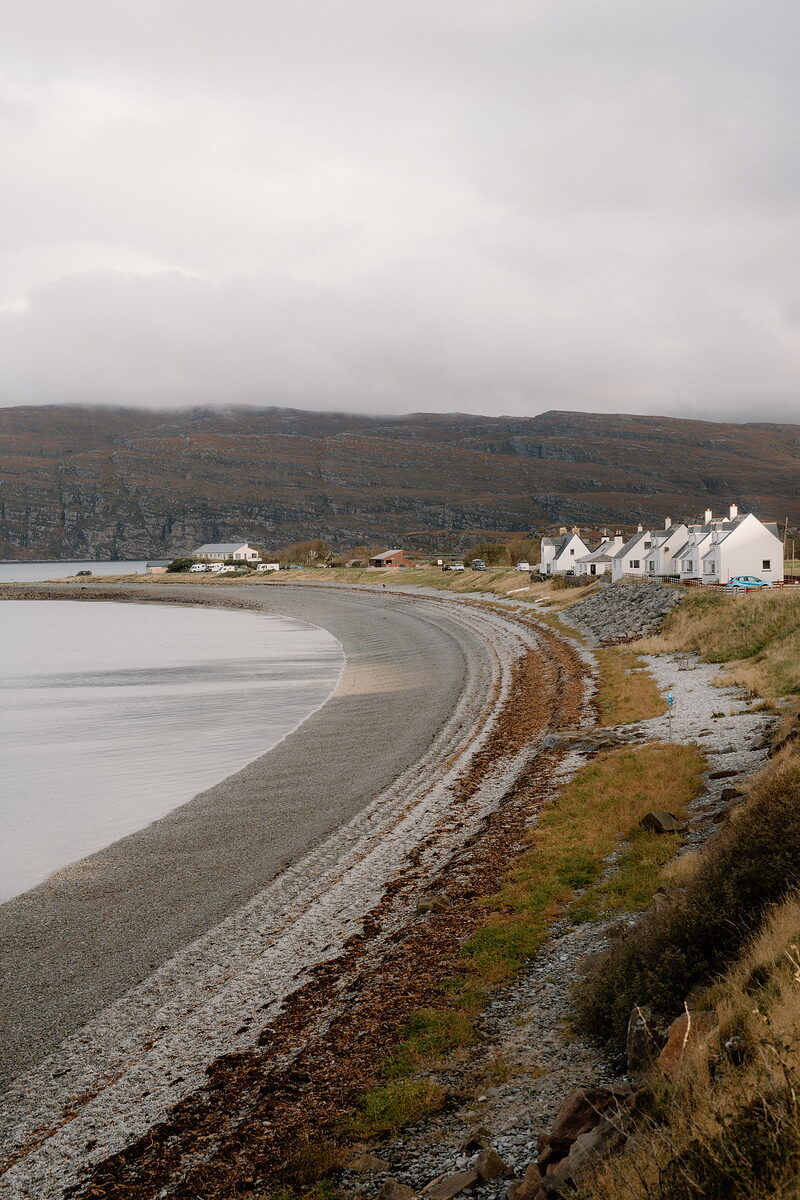 A row of whitewashed cottages lines a curving shingle beach beneath misty hills.