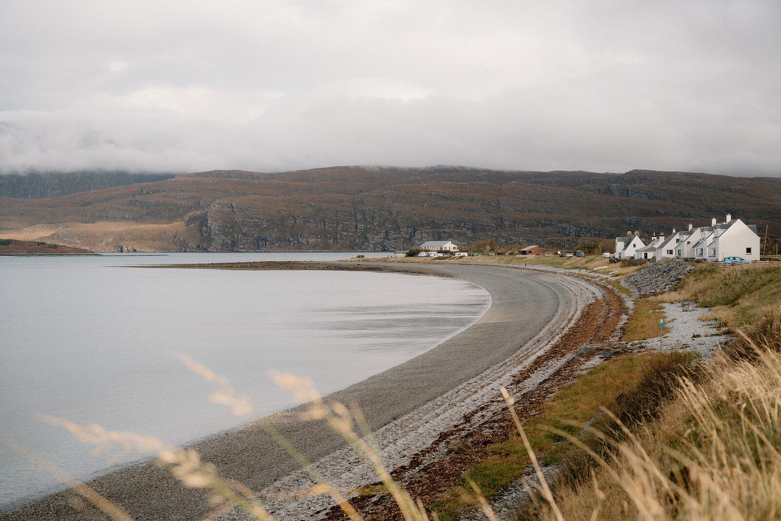 White cottages sit above a shingle beach curving around a calm sea loch beneath dark cliffs.