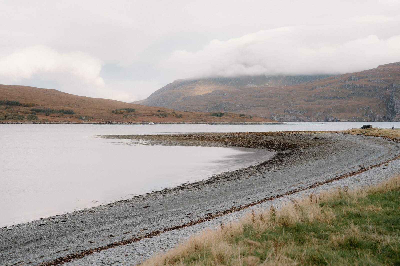 A curving shingle beach meets a calm loch beneath hills wrapped in low cloud.
