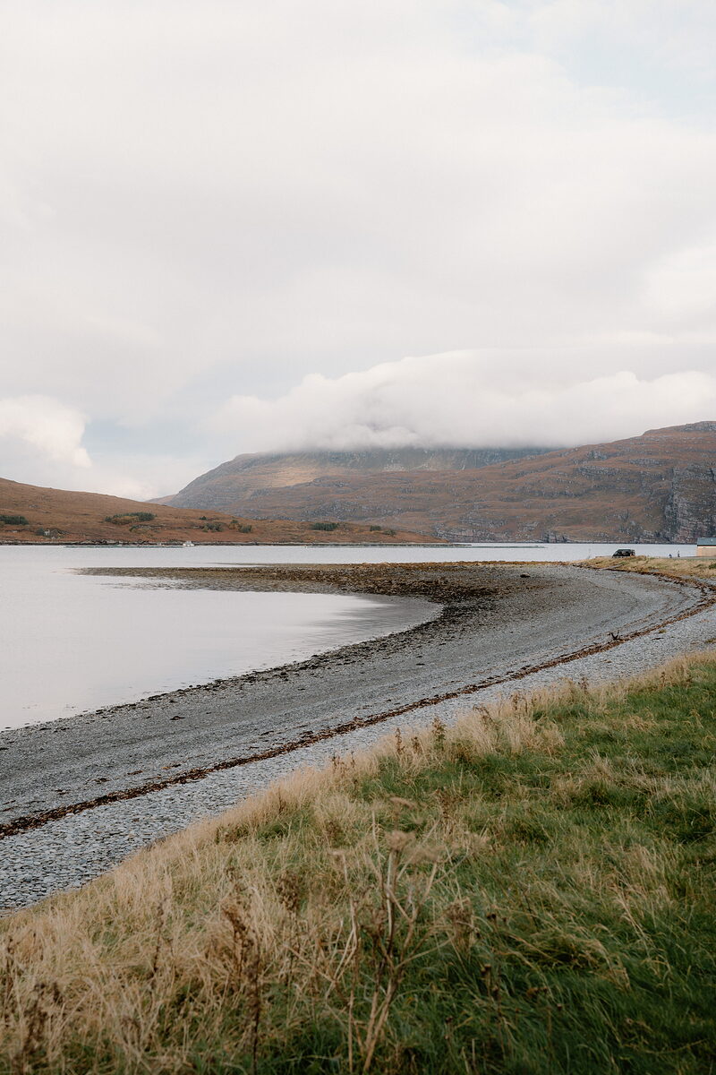 A gravel track follows a curving shingle beach beside a calm loch beneath misty hills.