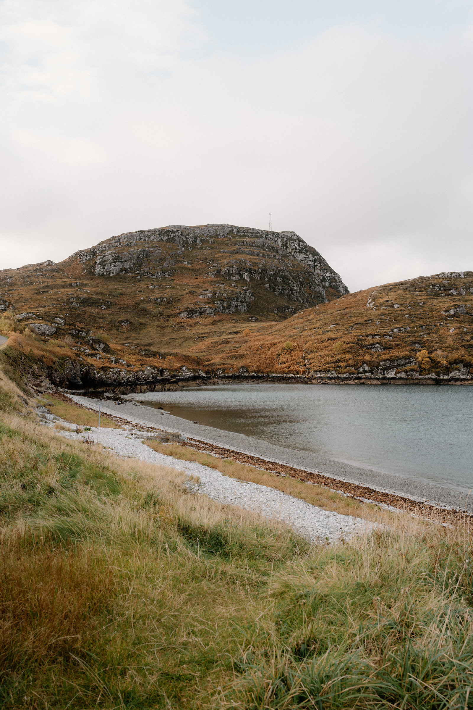 A curving shingle beach meets a calm sea loch beneath a rounded, rocky hill in Ross and Cromarty.