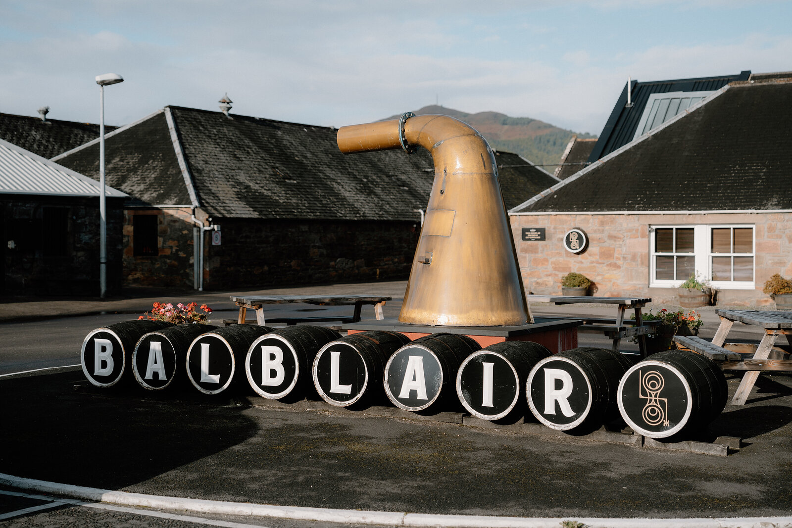 A copper still neck rises behind barrels spelling 'BALBLAIR' outside the distillery's buildings.