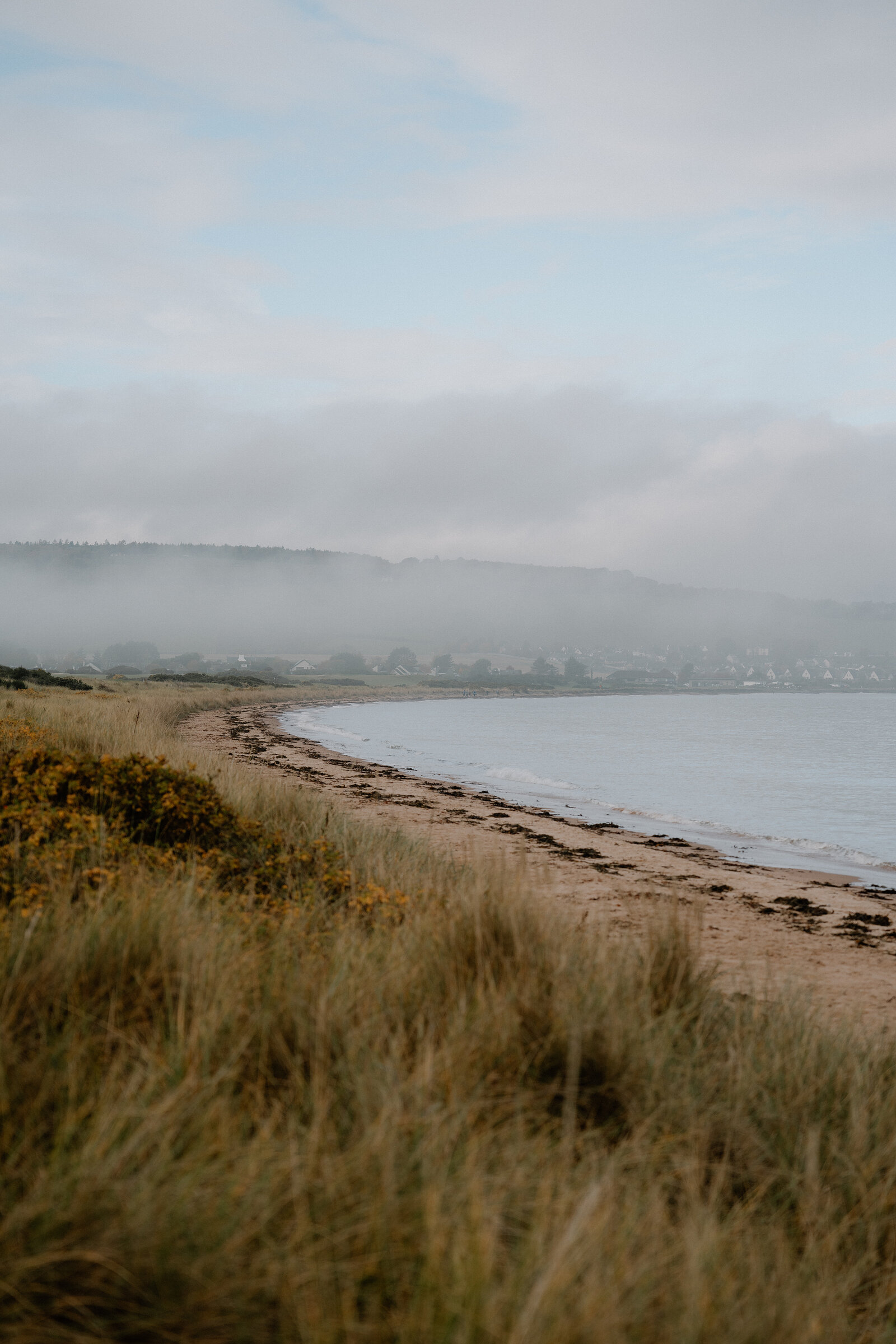 A sandy beach curves beneath dune grass toward a misty village in Ross and Cromarty.