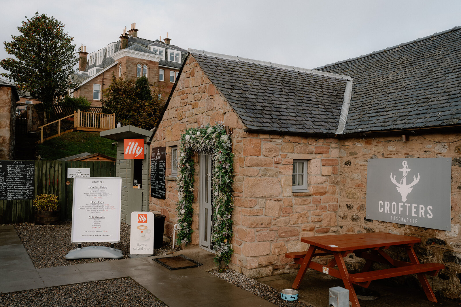 A stone building strung with fairy lights and greenery houses Crofters, its anchor sign beside a picnic table in Ross and Cromarty.