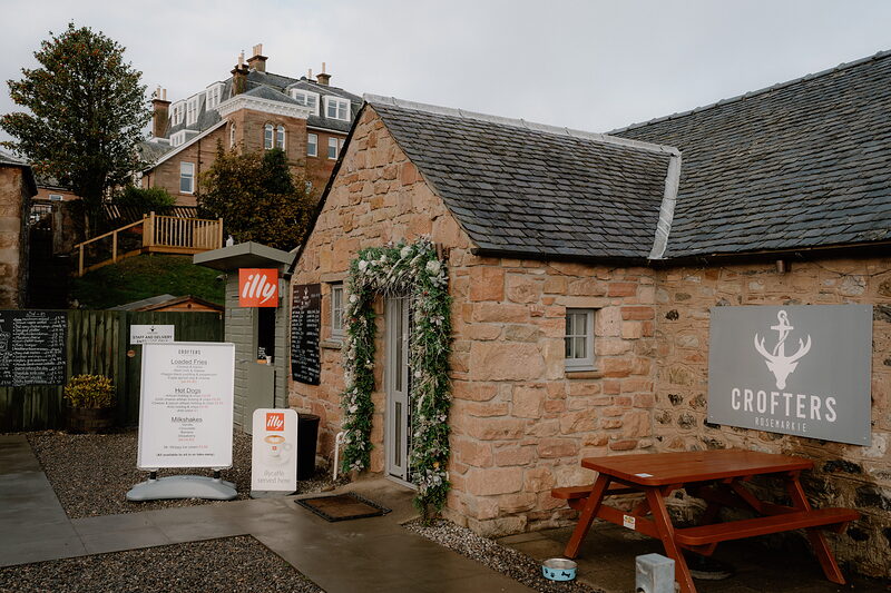 A stone building strung with fairy lights and greenery houses Crofters, its anchor sign beside a picnic table in Ross and Cromarty.