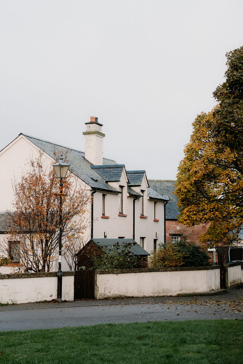 A white house with dormer windows and a tall chimney stands behind a lamppost and autumn tree in Cromarty.