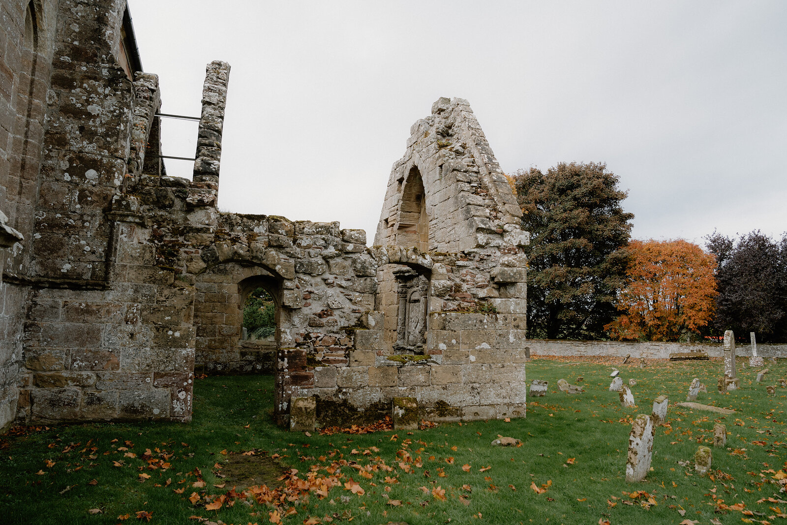 A gabled ruin with a pointed window stands beside an autumn tree in a Ross and Cromarty churchyard.