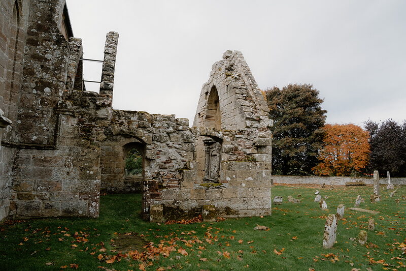 A gabled ruin with a pointed window stands beside an autumn tree in a Ross and Cromarty churchyard.