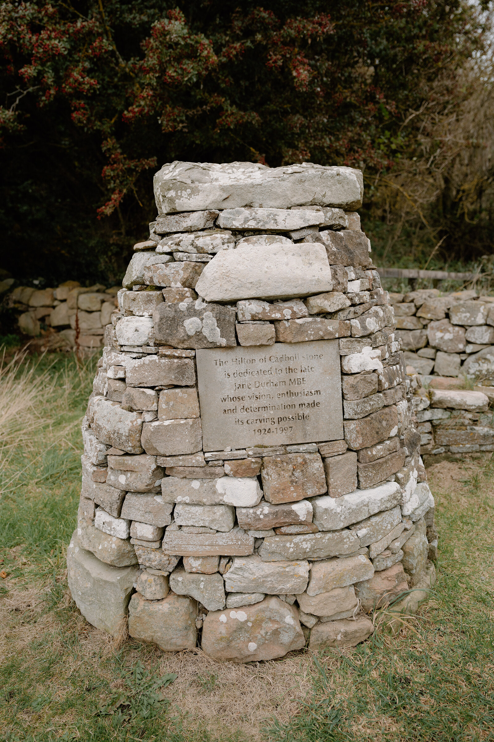 A conical stone cairn bears a weathered dedication plaque in a Ross and Cromarty garden.