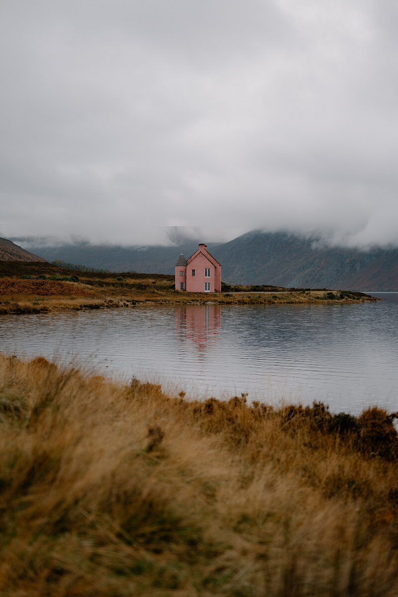 A pink cottage with a stone chimney stands alone on a loch shore in Ross and Cromarty, its reflection in the still water.
