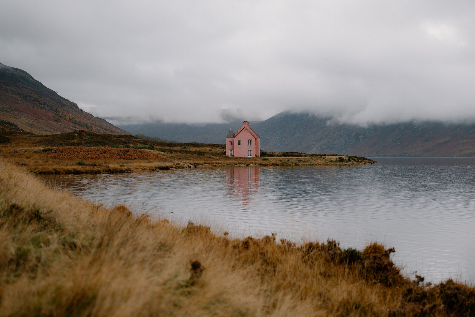 A small pink cottage sits isolated on a loch shore beneath layered mist and hills in Ross and Cromarty.