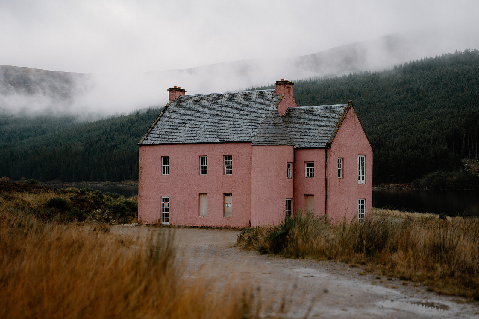 A pink house with chimneys and a corner turret stands by a gravel track against misty forested hills in Ross and Cromarty.