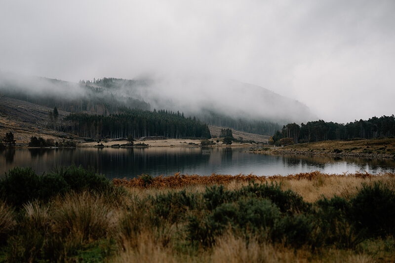 Mist rolls down a forested hillside above a still loch in Ross and Cromarty.