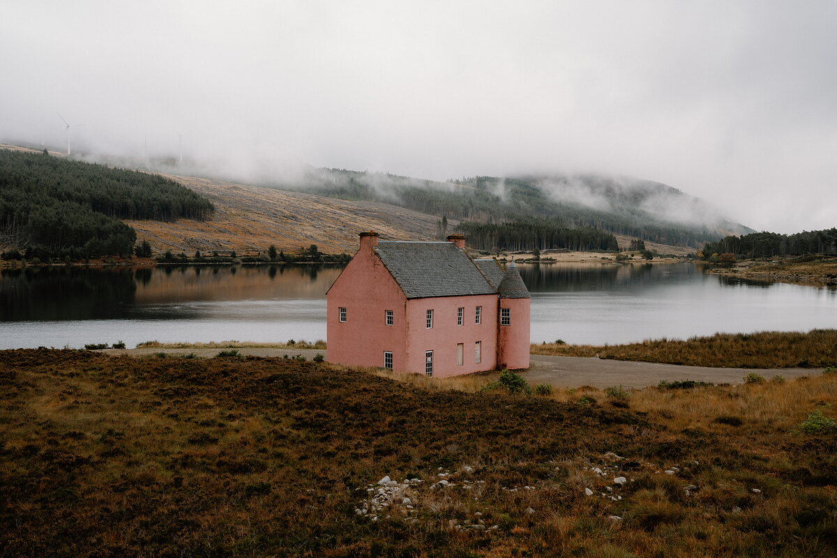 A pink mansion sits beside a calm loch in Ross and Cromarty, mist clinging to the forested hills beyond.