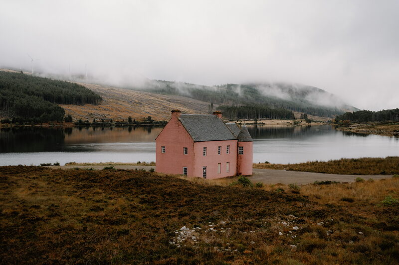 A pink mansion sits beside a calm loch in Ross and Cromarty, mist clinging to the forested hills beyond.